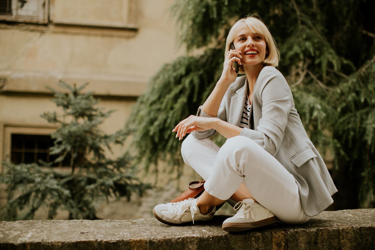 Femme souriante au téléphone, assise sur un muret urbain, en baskets compensées et tenue décontractée.