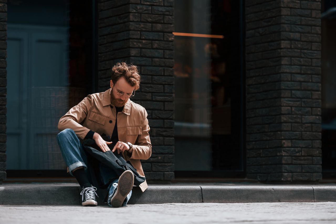 Homme stylé assis sur le trottoir, portant un manteau camel, jean brut et baskets bordeaux.